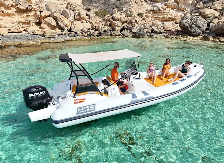 A white inflatable boat with a Suzuki outboard motor, labeled 'OROS MARINE', on clear turquoise water with rocky cliffs in the background. 3