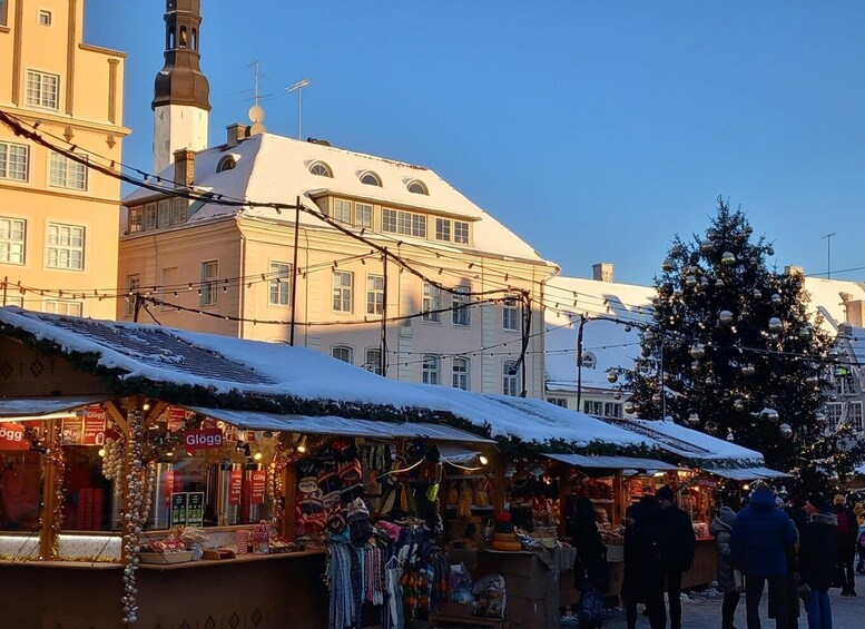 A snowy outdoor market with stalls selling various goods, a Christmas tree, and historic buildings in the background. 1