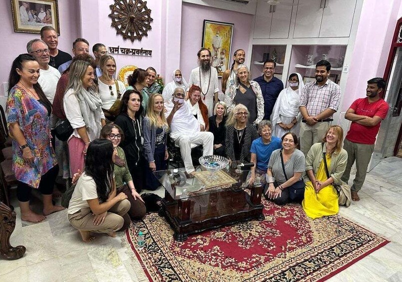 A group of people gathered in a room with a patterned carpet, a glass-top table, and a wall with a decorative item. 3