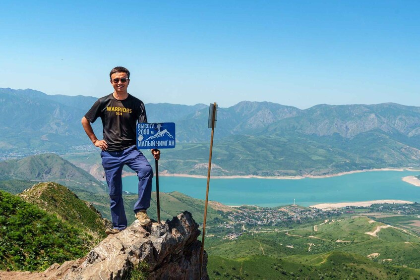 A person standing on a rocky outcrop with a sign indicating a mountain peak at 2099 meters. 1