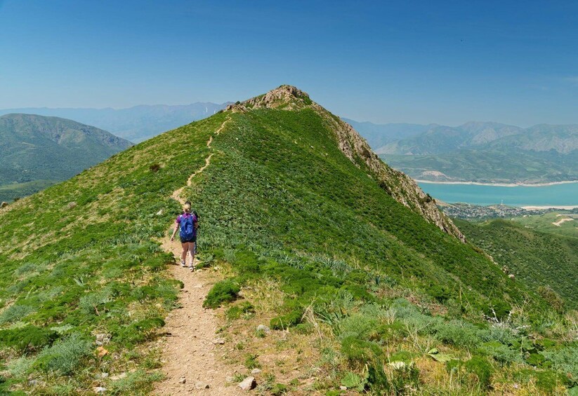 A hiker on a dirt trail on a green hill with a view of distant mountains and a beach. 2