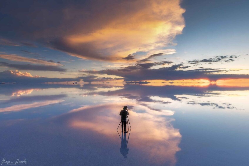 A person standing on a reflective surface with a camera, surrounded by a vast sky with clouds. 1