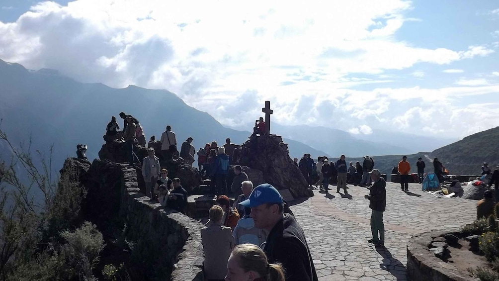 A group of people at a mountainous viewpoint with a cross monument. 1