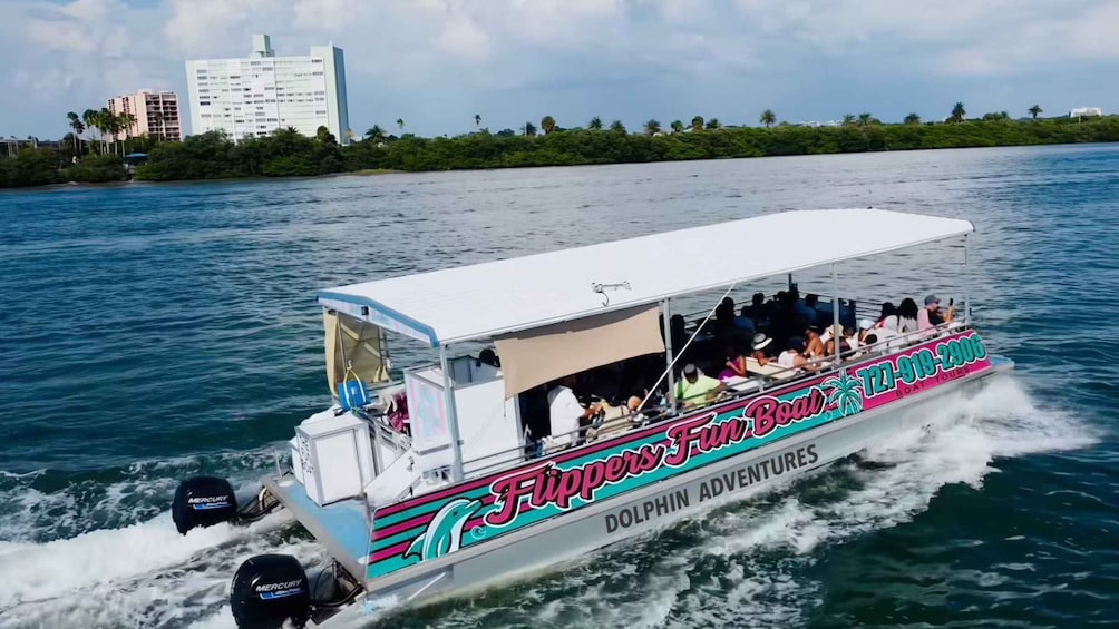 A boat named 'Fun Boat Tours' with 'Dolphin Adventures' written on the side, moving through water with a cityscape and greenery in the background. 2