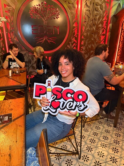 A person holding a sign with 'Pisco Lovers' in a restaurant with a decorative wall and a circular sign with 'EDEN' on it. 2