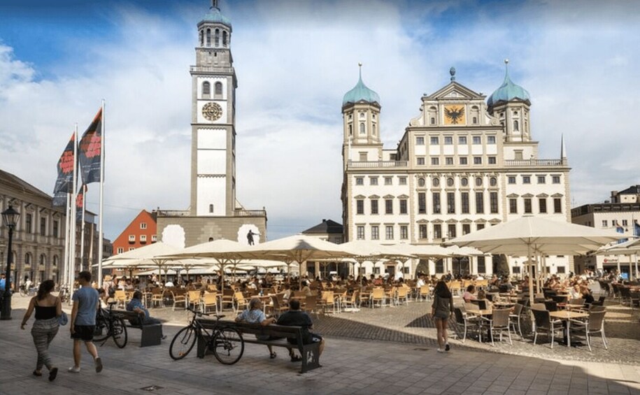 A public square with outdoor seating, a clock tower, and a large building with domed roofs. 1
