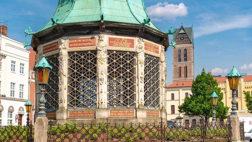 A historical building with ornate metalwork and inscriptions, surrounded by a fence and other architecture. 2