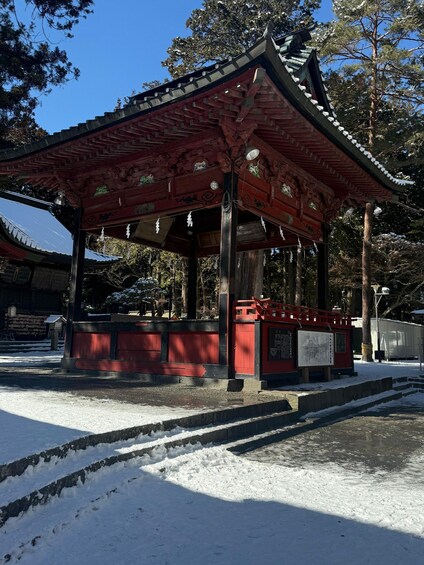 A traditional Japanese pavilion with a red roof and intricate woodwork, surrounded by snow-covered ground and trees. 2