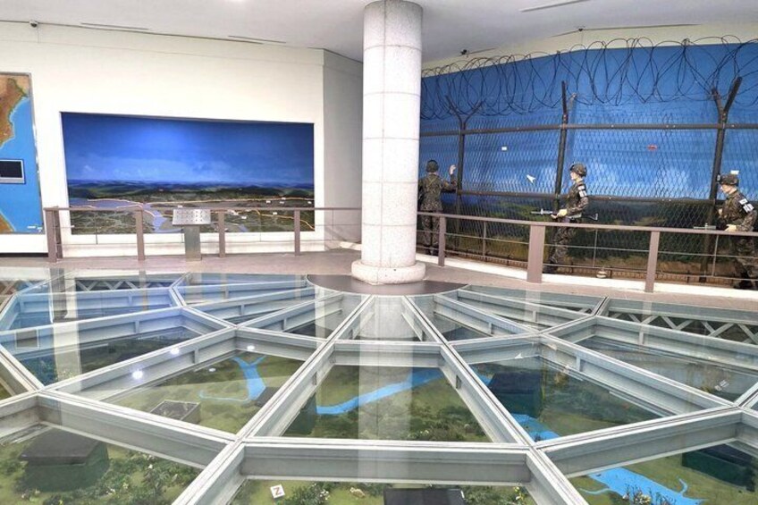 A museum interior with a glass floor, a large blue painting, and a fence with barbed wire. 5