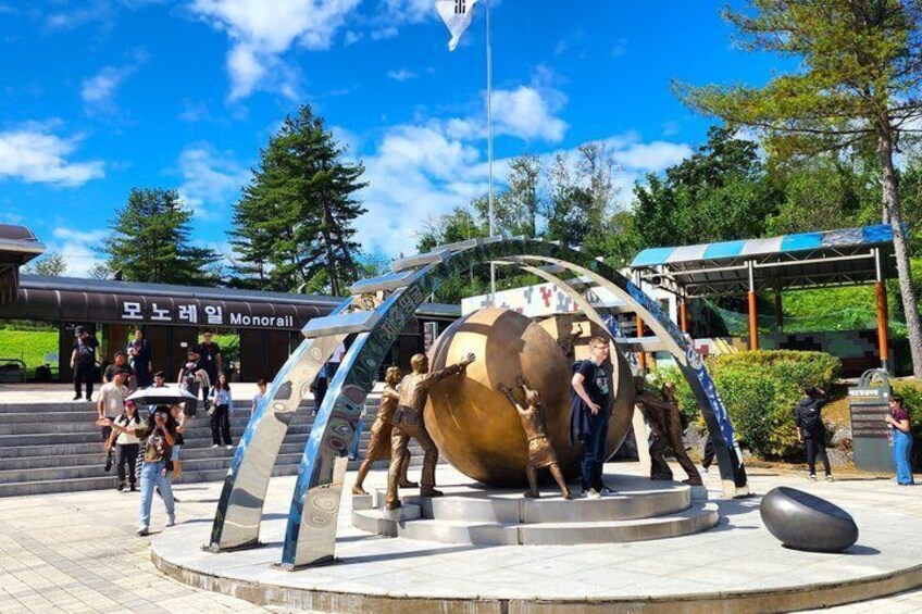 A large metallic sculpture with a circular base, surrounded by a paved area with steps leading up to it. 1