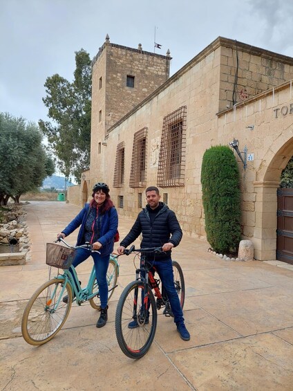 Two people on bicycles in front of a historic stone building with barred windows and a sign reading 'TOR.' 1