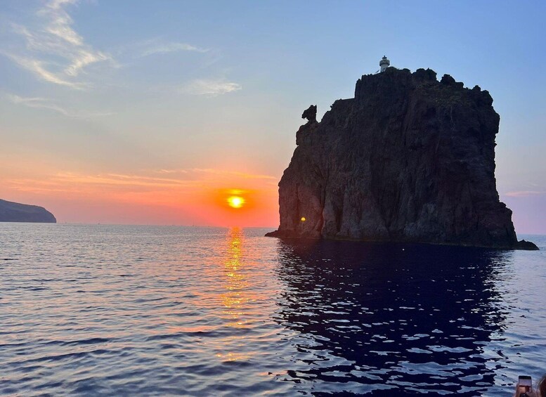A sunset over a calm sea with a rocky outcrop and a lighthouse. 2