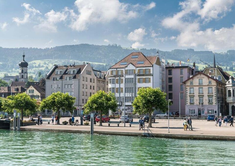 A riverside promenade with buildings, trees, and a distant hill. 3