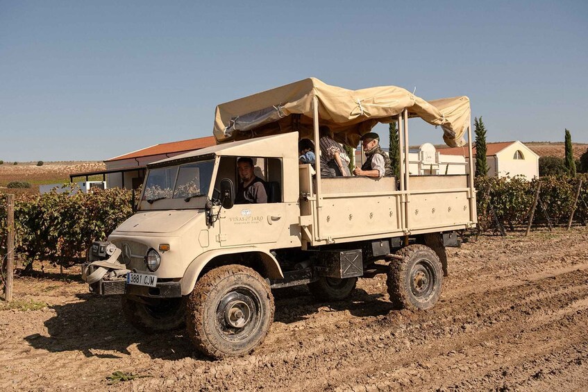 A beige off-road vehicle with a canopy, parked on a dirt road in a rural area with vineyards and houses in the background. 5