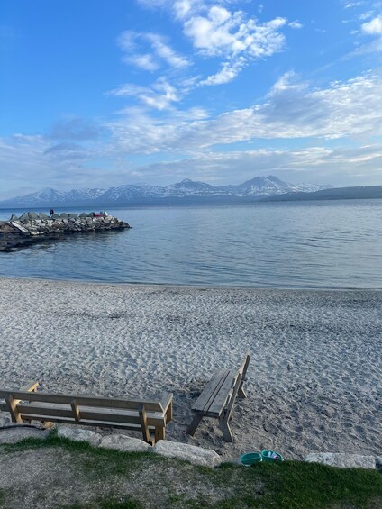 A pebble beach with a wooden bench and picnic table, a rocky breakwater, calm water, and snow-capped mountains in the distance. 1