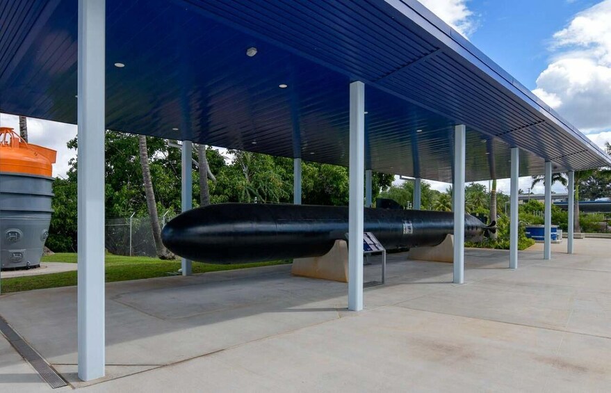 A covered outdoor area with a large black cylindrical object, possibly a sculpture, under a blue roof supported by white pillars. 5