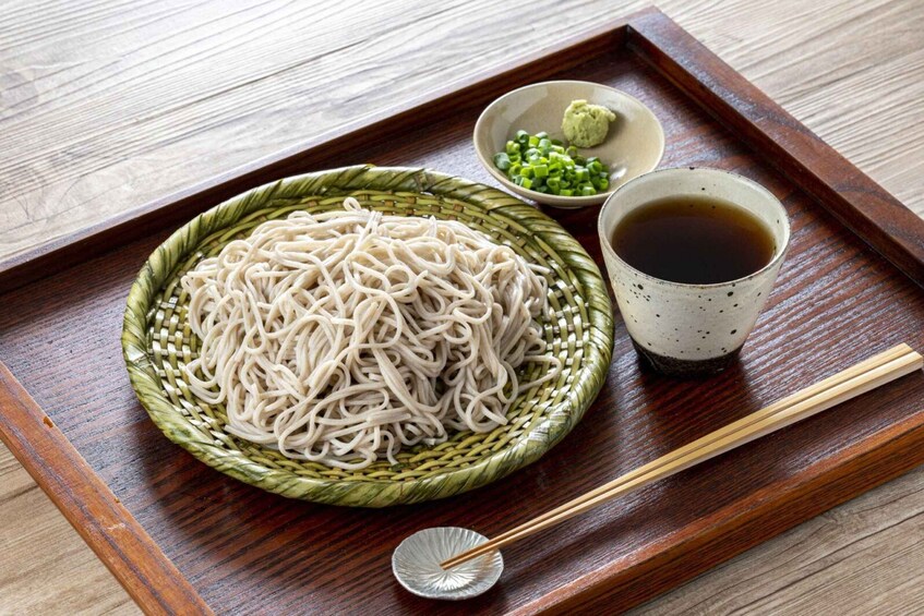 A plate of noodles with chopsticks, a small bowl of green onions, a bowl of sauce, and a cup of tea on a wooden tray. 2