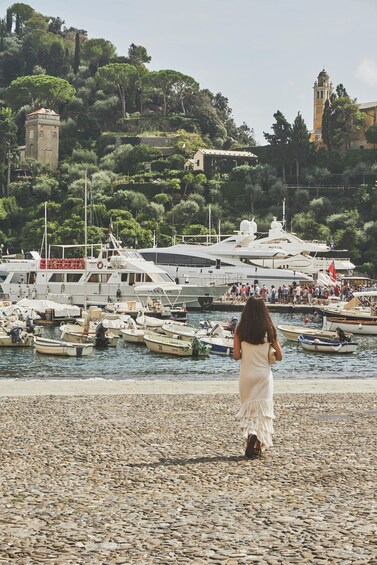A rocky beach with a woman in a white dress walking away from the camera, boats docked in the water, and a tower on a hill in the background. 5