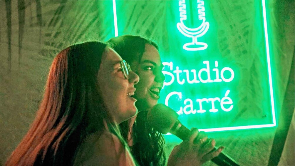 Two people holding a microphone with a neon sign in the background that reads 'Studio Carré.' 1