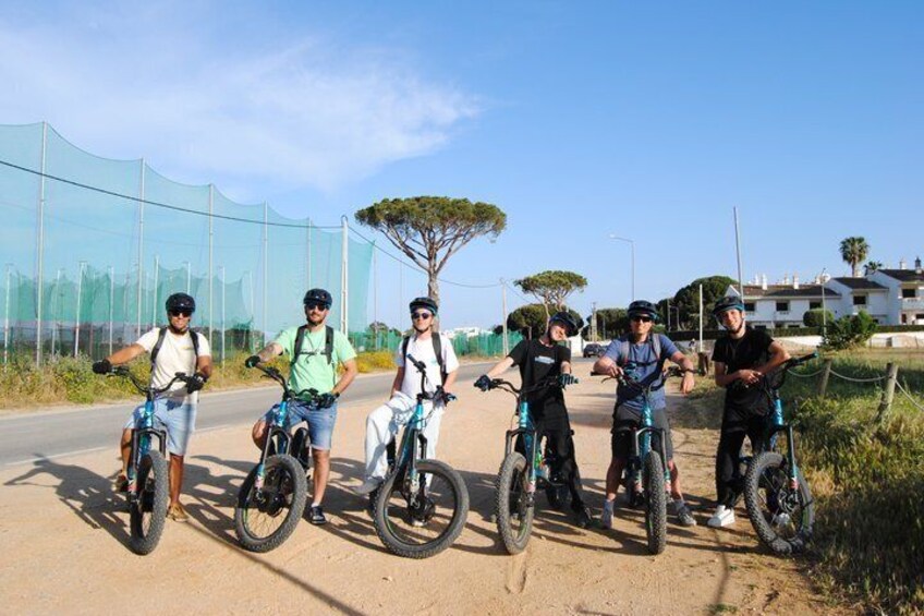 A group of cyclists on a paved road with a fence and trees in the background. 4