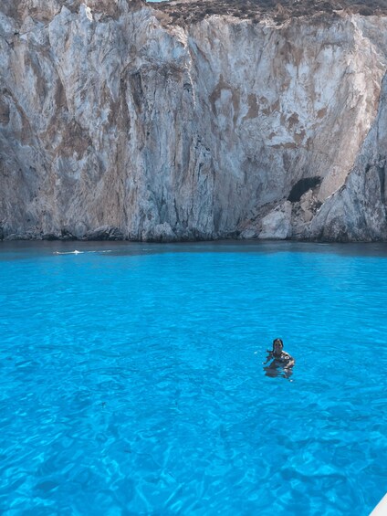 A person swimming in clear blue water with a rocky cliff in the background. 2