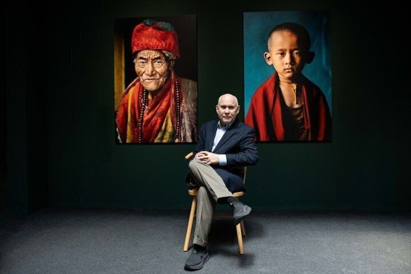 A man seated in a chair with two framed portraits on the wall behind him. 1