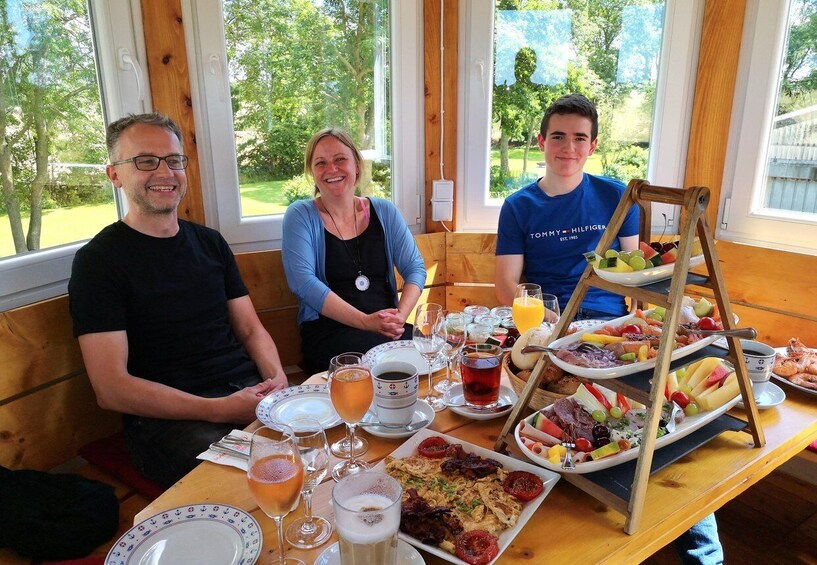 Three people seated at a table with a variety of food and drinks, including a fruit and cheese board, in a room with large windows. 4
