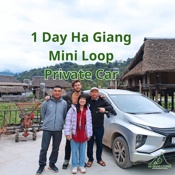 A group of people posing in front of a silver car with a thatched-roof building in the background. 1