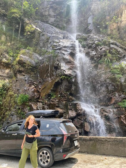 A woman standing in front of a waterfall with a car parked nearby. 5