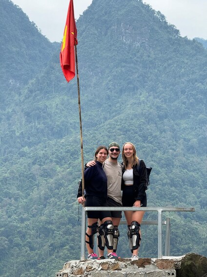 Three people standing on a rocky ledge with a forested mountain in the background. 3
