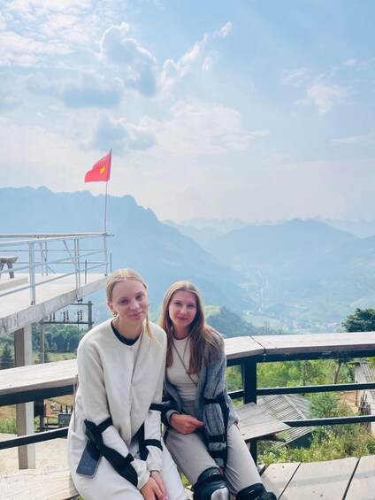 Two people sitting on a wooden bench with a mountainous landscape and a flag in the background. 4