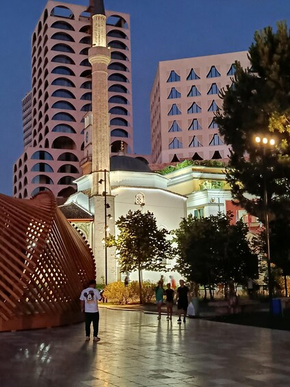 A modern building with a unique facade, a tall tower, and a plaza with people and trees. 2