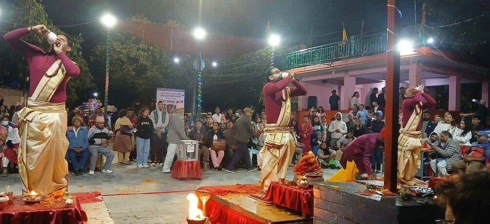 A cultural performance with performers in traditional attire on a stage, an audience in the background, and a decorated outdoor setting. 1