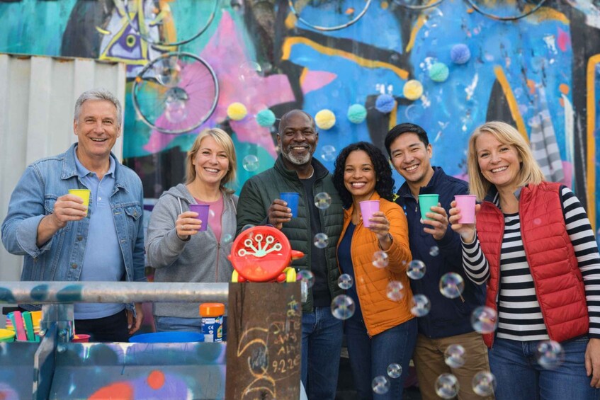 A group of people standing in front of a colorful mural with a bubble machine in the foreground. 1