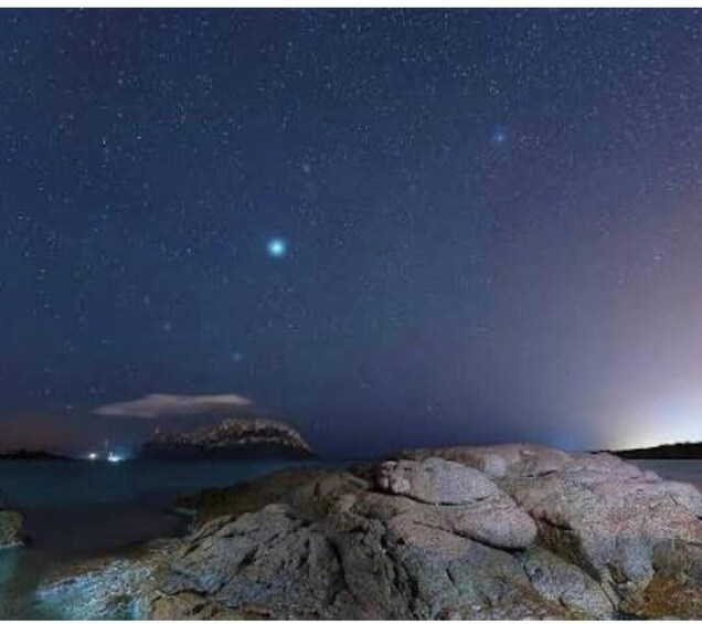 A starry night sky over a rocky coastline with a visible island in the distance. 4
