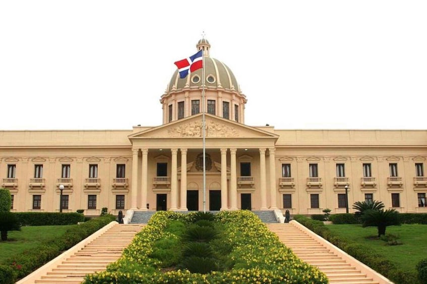 A large neoclassical building with a central dome, flag, and formal garden. 1