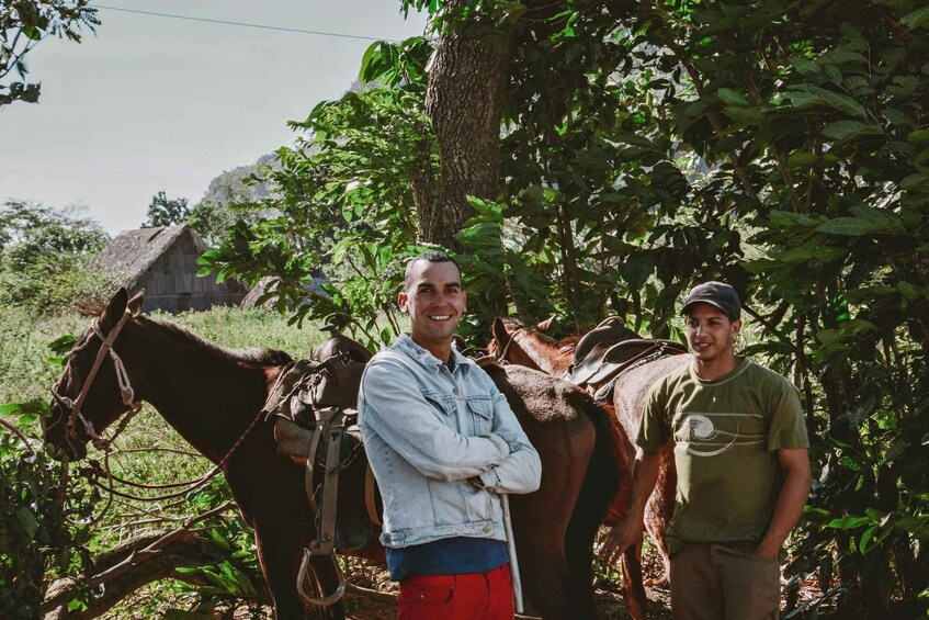 Two men standing with horses in a rural setting with trees and a thatched-roof building in the background. 2