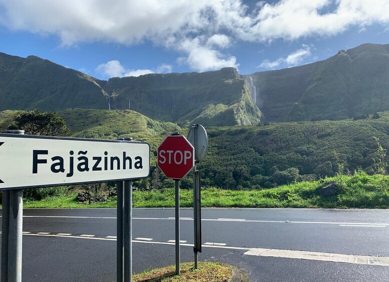 A road sign with 'Fajãzinha' and a STOP sign against a backdrop of lush green hills and a waterfall. 5