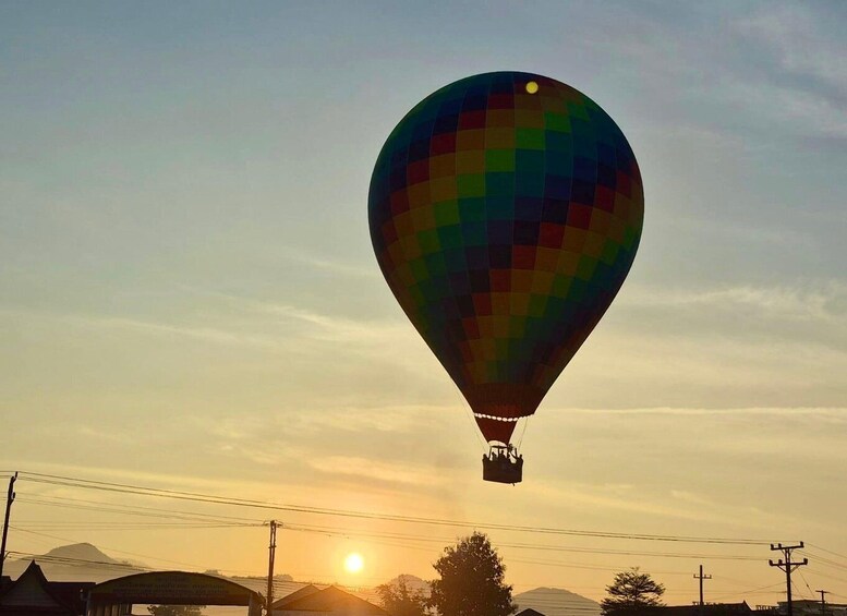 A hot air balloon with a colorful pattern, flying above a landscape with buildings and power lines, during sunset. 3