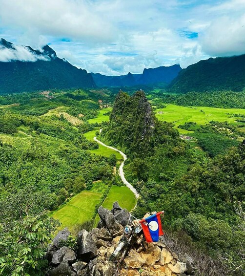 A mountainous landscape with lush greenery, a winding path, and a flag on a rock. 1