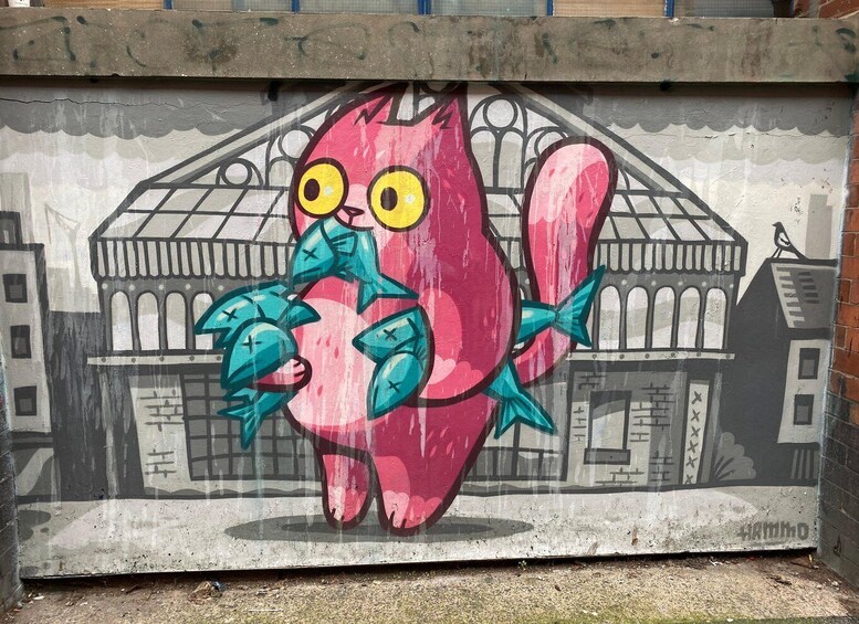 A mural of a pink bird with blue wings and yellow eyes holding a blue fish, set against a backdrop of a building with a tiled roof. 2
