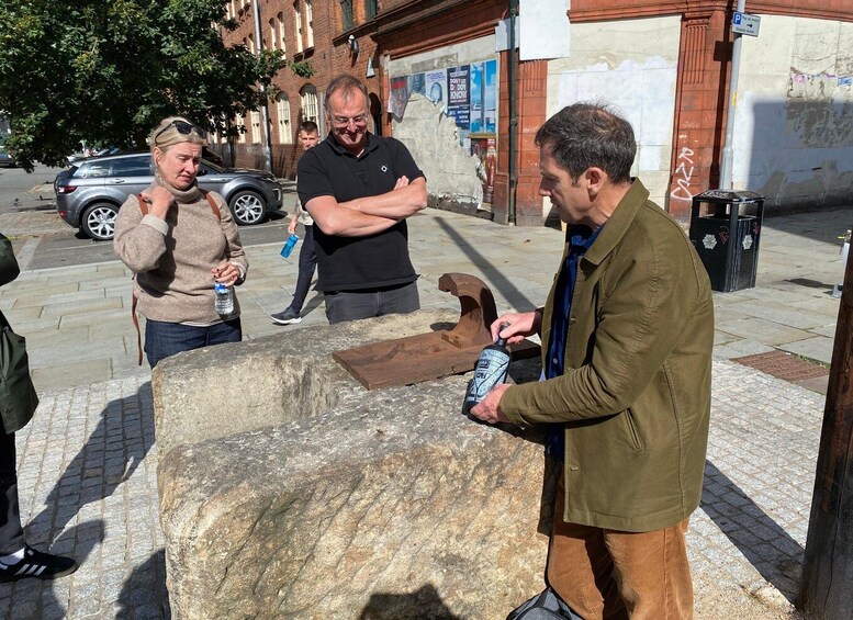 A group of people standing around a large stone slab outdoors, with a car and buildings in the background. 1