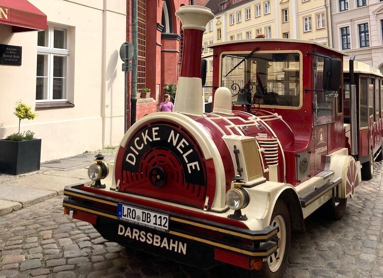 A vintage red and cream train with 'DICKE NELE' written on the front, parked on a cobblestone street. 5