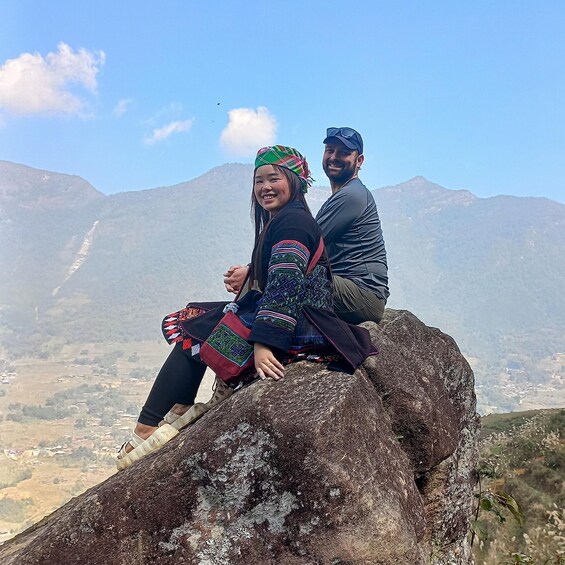 Two people sitting on a rock with a mountainous landscape in the background. 1