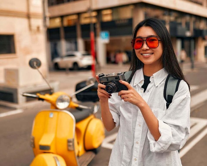 A person holding a camera, standing next to a yellow scooter on a city street. 3