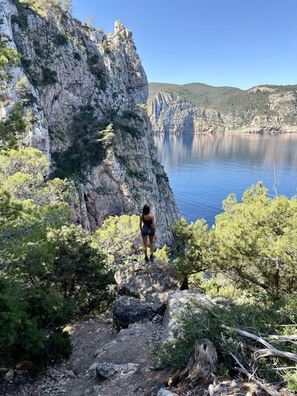 A person standing on a rocky outcrop overlooking a calm lake with cliffs in the background. 2