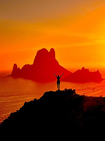 A person standing on a cliff with arms raised, silhouetted against a sunset with red and orange hues, with mountains in the background. 1
