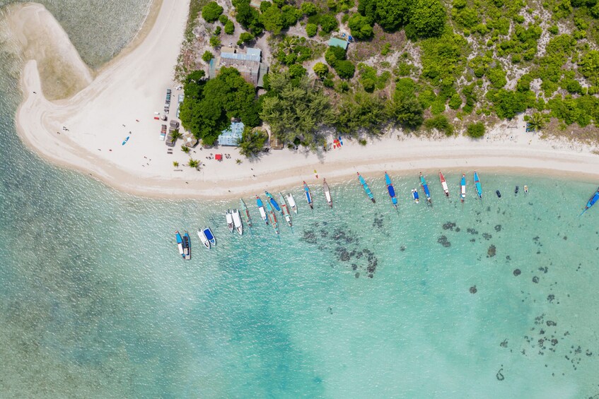 An aerial view of a beach with a row of boats anchored in the water, a sandy shoreline, and a backdrop of greenery and buildings. 2