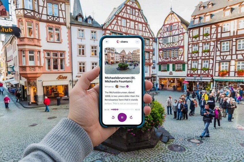 A hand holding a smartphone displaying a location named Michaelisbrunnen (St. Gutenberg) in a European town square with traditional half-timbered buildings. 1