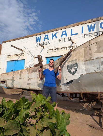 A man holding a wooden plank in front of a rusted ship with 'WAKALIWO' and 'RAMON FILM PROD' written on it. 4